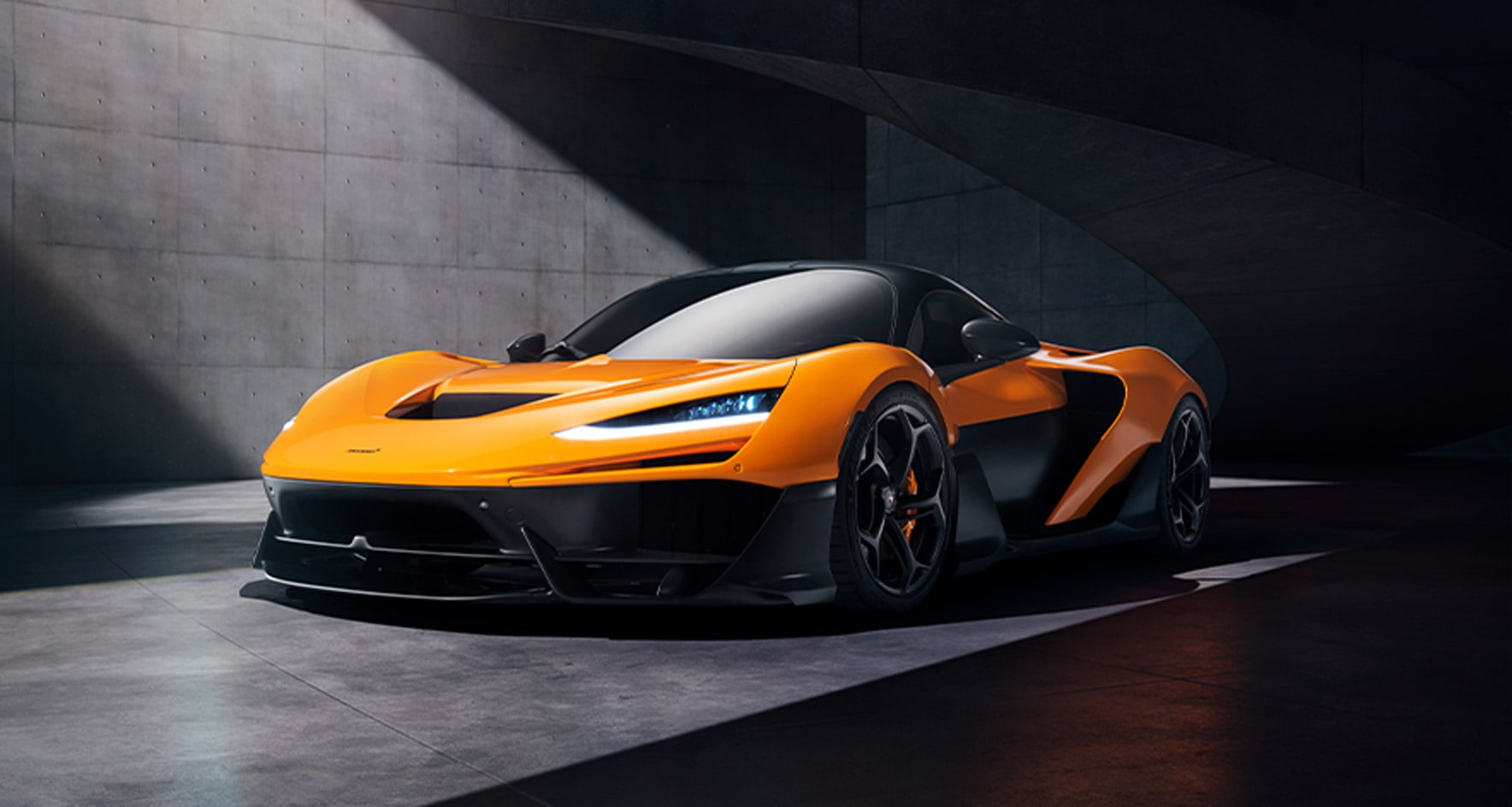 A front three-quarter view of the orange McLaren W1 parked in a modern, concrete garage, emphasizing its sleek headlights and aerodynamic front splitter.
