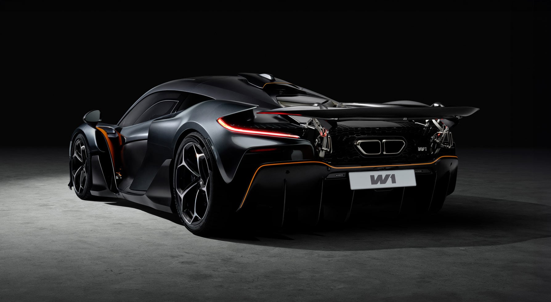 A rear view of the black McLaren W1 illuminated by a spotlight, highlighting its complex rear end with active aero and prominent exhaust outlets.