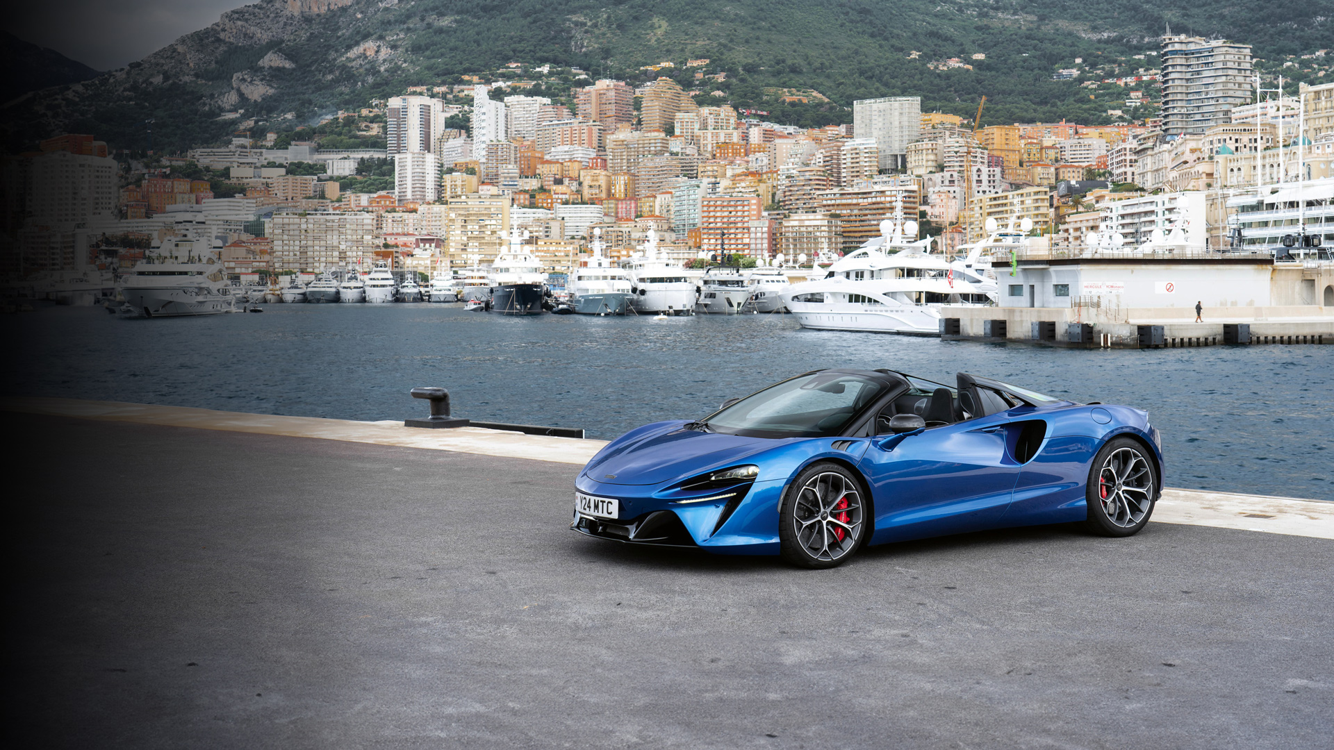 The blue McLaren Artura Spider, an open-top hybrid supercar, parked dockside in Monaco’s Port Hercule. The image captures the lightweight performance machine against a backdrop of luxury yachts and the iconic Monte Carlo landscape, emphasizing exclusivity and dynamic driving capability.