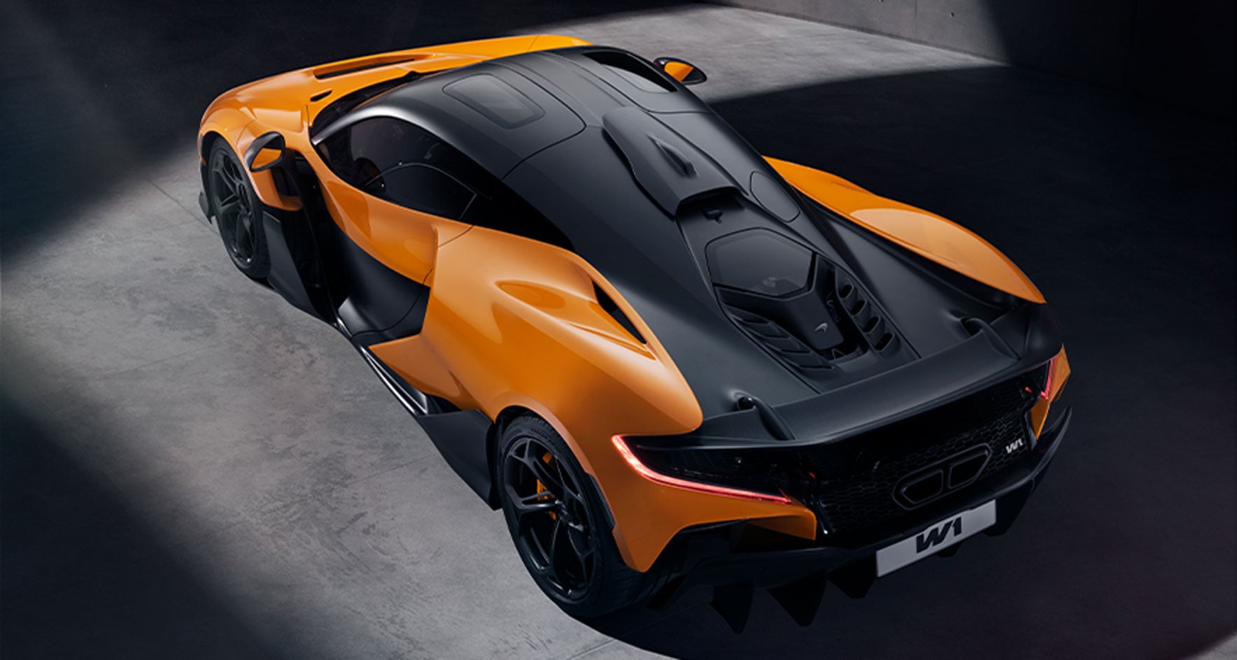 A high-angle, rear three-quarter view of the McLaren W1, a vibrant orange hypercar with a black roof and engine cover.
