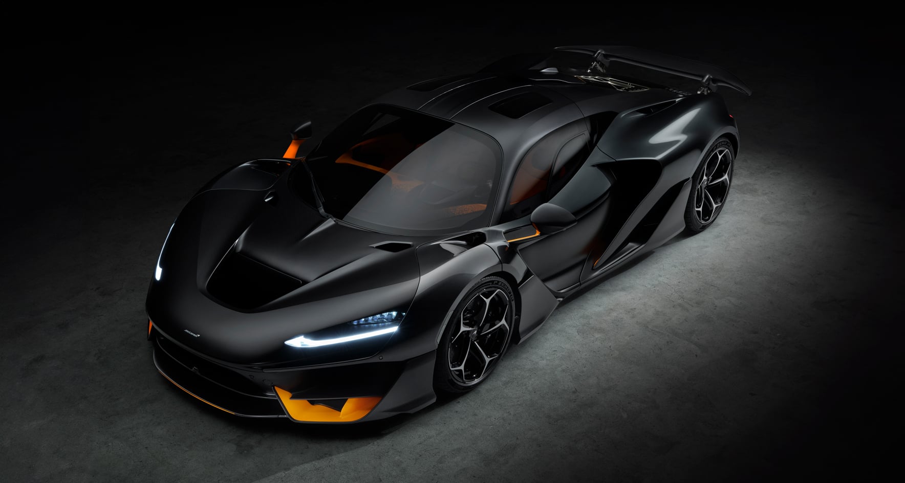 A high-angle, front-view shot of the McLaren W1, a sleek, matte black hypercar with sharp lines and subtle orange accents on the front splitter and brake calipers, parked in a dark, spotlighted setting.