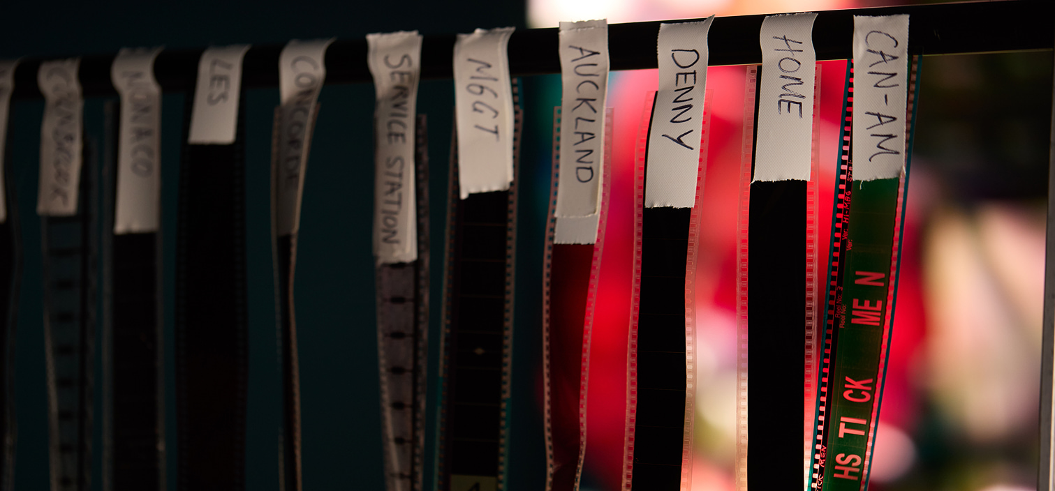 Rows of labeled film strips hanging vertically, each tagged with handwritten names like “Auckland,” “Denny,” and “Home” against a softly blurred background.