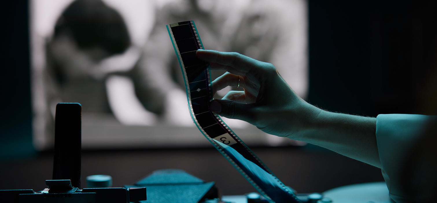 A hand holds a strip of photographic film up to the light while a blurred image appears in the background.
