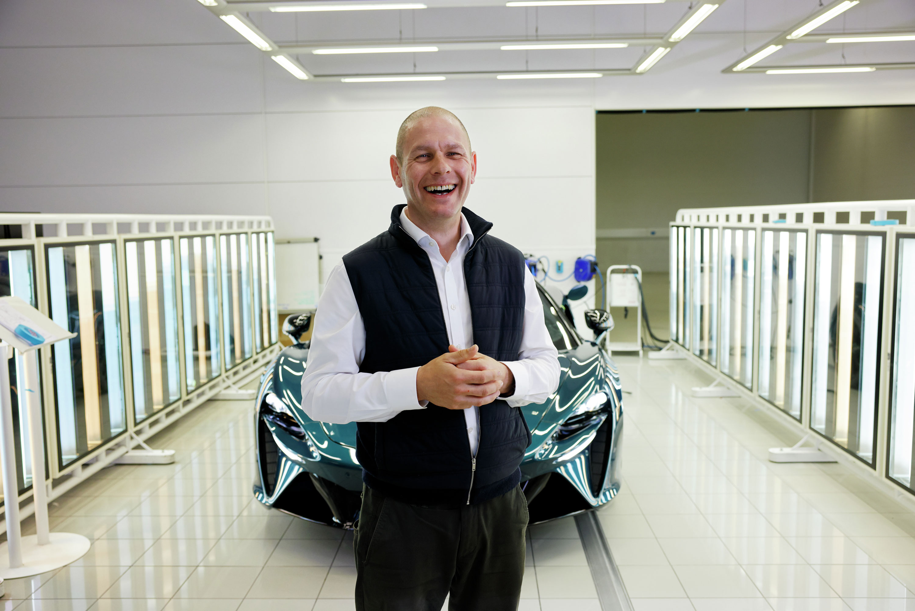 A laughing man standing in a McLaren factory with a teal Artura supercar in the background.